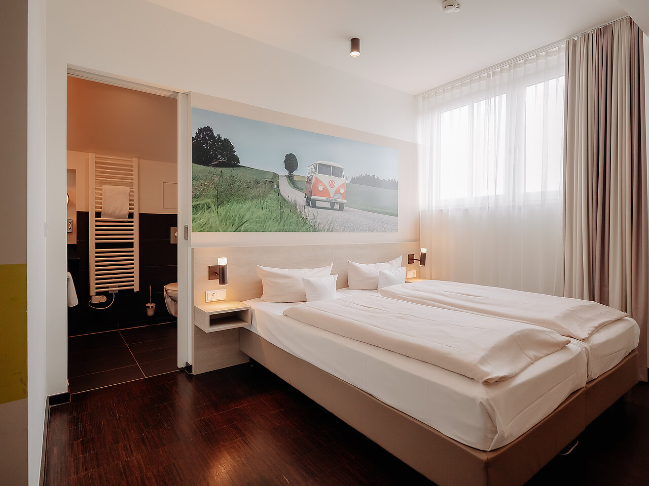 Doppelzimmer mit Bulli-Motiv im CrossINN Osnabrück Helles Doppelzimmer im CrossINN Osnabrück mit zwei Betten und großem Wandmotiv eines VW Bulli